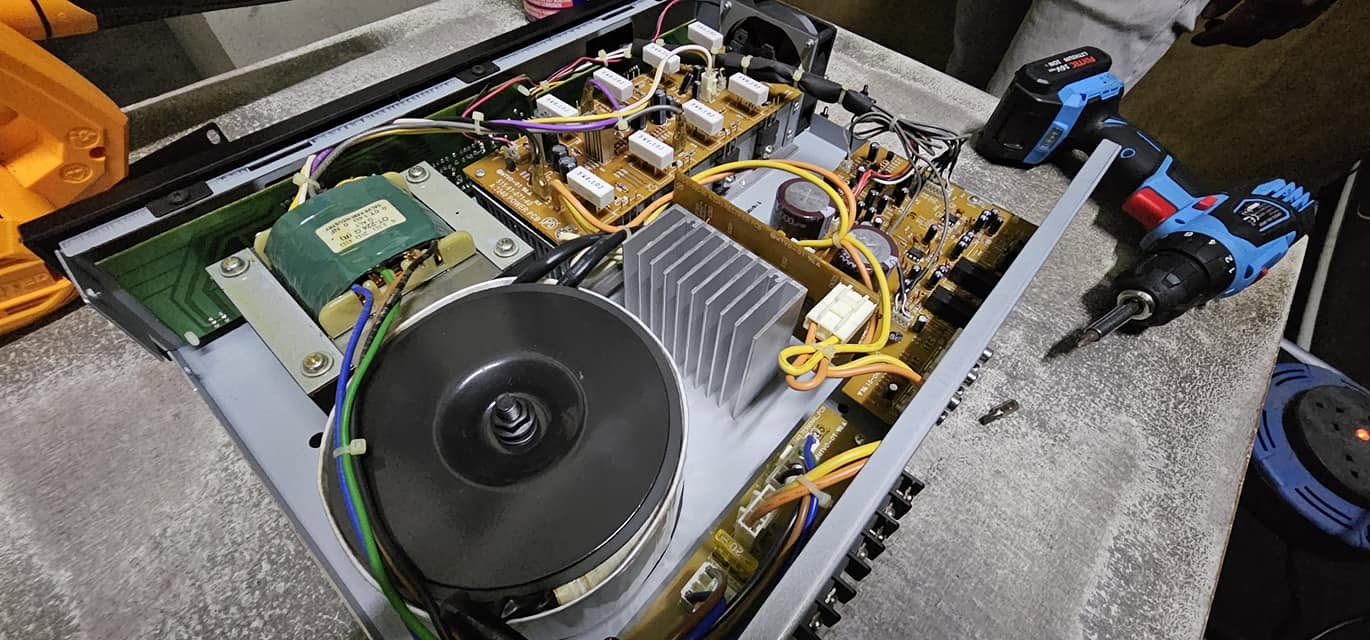 Open amplifier chassis on the workbench with toroidal transformer, heatsink, control boards and a cordless driver ready for repair